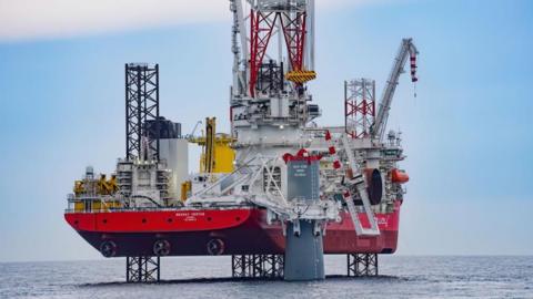 A close up image of Seaway Ventus is what is known as a 'jack up barge' and this image shows the vessel raised out of the sea on four legs. On the deck among the superstructure is a giant crane, which has a line attached and is installing the first turbine 'monopile' into the sea bed. This steel structure is grey, and tubular. There is a second crane and other infrastructure and equipment on the deck.