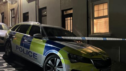 A police car is parked on a street at night-time. Police tape is hanging between the car and a hose. The houses are terraced.