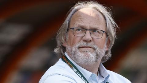 Tom Saintfiet in smart shirt and with lanyard round neck looks up while on the touchline