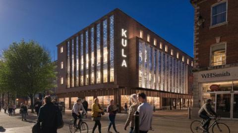 The outside of a hotel, which has the words 'Kula' written on it, showing the lights on at night. There are also pedestrians and cyclists passing by outside.
