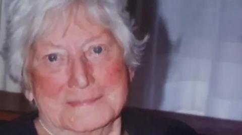 Janet Purkess in an old photo, looking at the camera and smiling slightly. She is older and has white hair.