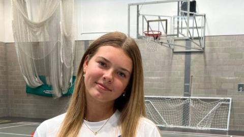 A medium close-up, eye-level photo shows a young woman (Vanessa) with long, light brown hair smiling faintly at the camera. She is standing in a sports hall. Behind her, a basketball hoop with a net and backboard is visible. She is wearing a white T-shirt with a small logo on the right side of her chest. The lighting is bright and uniform.