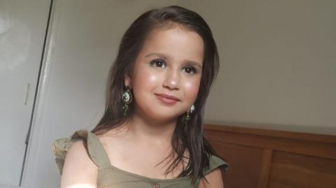 Sara Sharif, a 10-year-old girl, smiling as she sits on a bed while wearing a green dress