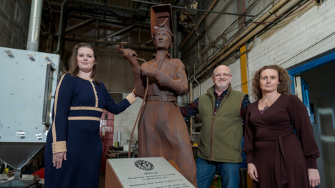 The statue of Molly. It is brown and made of corten steel. The figure holds a welding torch and is wearing an open visor so her face is revealed. To her left stands Dr Ashleigh Blackwood wearing a blue dress. To her right is Dr Ron Lawson wearing a green gilet and Dr Victoria Rafferty in a maroon dress.