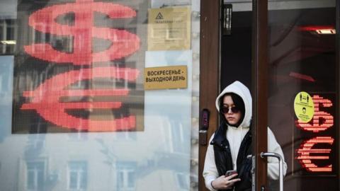 Woman in sunglasses and wearing a grey hooded sweatshirt leaves a Moscow money exchange