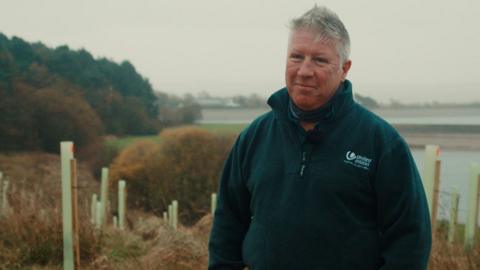 Woodland project officer Michael Bullivant standing by the edge of the reservoir alongside the saplings. The sky is plain grey and he is wearing a dark green fleece and smiling. He has short grey hair.