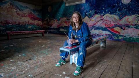 Emily Powell photographed in her barn. She is wearing a blue jumpsuit that is covered in paint. She is holding a jar with water and brushes inside while she is sat on a bench. She is also surrounded by her artwork on the walls.