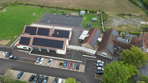 An aerial image of school buildings with a number of large solar panels attached to its roofs. There is also a field, car park and football pitch in the vicinity of the buildings.