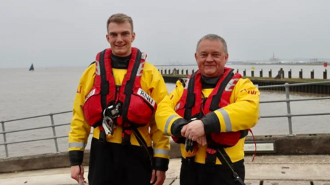 Harry standing with his Dad, both in RNLI yellow uniform and wearing red life jackets, on the launch ramp at Portishead Lifeboat Station