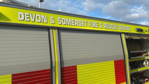 The side of a Devon and Somerset Fire and Rescue Service fire engine. The vehicle has red and yellow hi-vis stickers on its exterior. It has two closed hatches. One hatch is open exposing fire equipment.