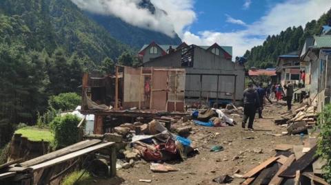 People walk down a dirt road amid rubble and destroyed buildings beside a partially collapsed hillside