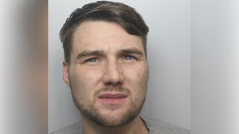 A mugshot of a man with short brown hair, stubble around his chin and a grey top.