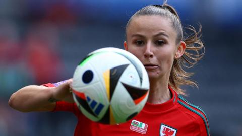 Hannah Cain shown behind a football which is photographed in the air in front of her. Her right arm is folded behind the ball at the left of the picture. She is wearing red Welsh football kit.