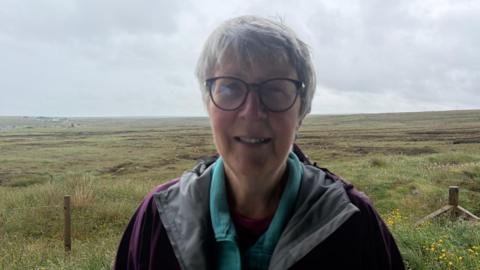 Boireannach le falt goirid, liath agus speuclairean na seasamh air a' mhòinteach.