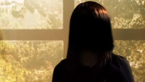 "Sarah", with her back towards the camera, is silhouetted against a brightly lit window. She has dark hair and is wearing a black top. 

