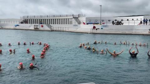 A number of swimmers with pink and yellow swim caps float with their heads above the water in a large open saltwater pool, with a white surround and some white-painted concrete changing booths on the far side from the camera.