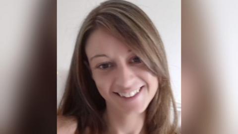 A portrait image of a woman with long brown hair smiling while in front of a white wall.
