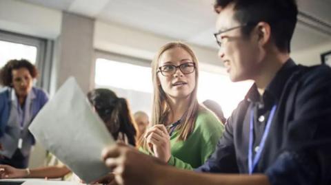 Two students wearing glasses and jumpers sit at desks in a classroom, with a teacher and other students in the background.