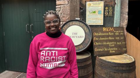 Janett Walker in pink jumper which says Anti Racist Cumbria. She has large colourful glasses and is smiling at the camera. She is standing by barrels and a wooden sign listing different types of Jamaican rum.