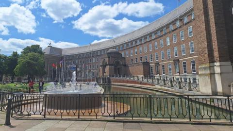 City Hall in Bristol, a red brick crescent-shaped building with five stories, a grey pitched roof and a fountain in front.