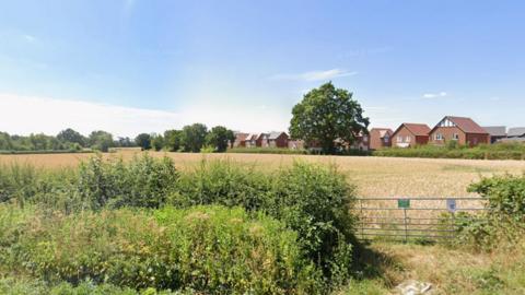 Site earmarked for 91 new homes. It is a large field with a gate next to a hedgerow. Homes can be seen at the far side of the field against a blue sky.