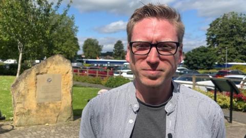 Lib Dem Councillor Dan Boatright-Greene is standing outside in the sunshine, wearing a stripy open-necked shirt.