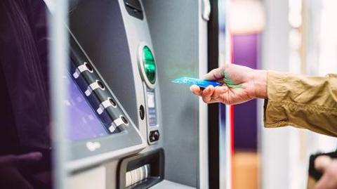 A person reaches up to a ATM machine with their blue bank card in their hand.