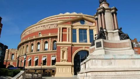 Exterior shot of Royal Albert Hall and and statue with blue skies behind.