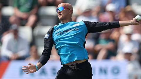 Fateh Singh is about to complete his bowling action in a white-ball game for Worcestershire. He is wearing black trousers and a top with black sleeves and light blue front. He is also wearing sunglasses