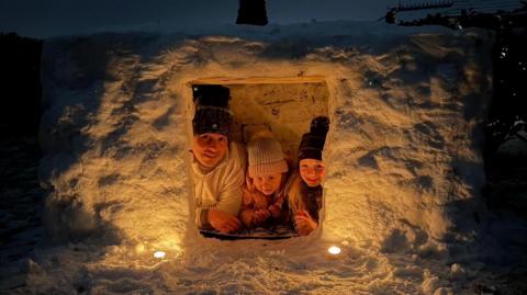 Three people lie on their fronts, with their faces up looking into the camera. They are inside a small white structure made of snow. Inside the on the left is a man wearing a white jumper and black hat. A small girl has on a pink coat and grey and pink hat. The girl to the right wears a black hat. There are two small lights at the entrance that illuminate the structure. Its dark outside