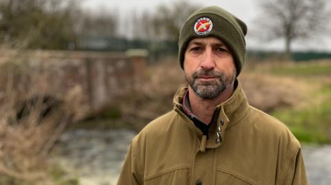 Matt Arnold frowns as he looks towards the camera. He has a dark, short beard. He is wearing a green woollen hat with a red and yellow badge on the front and a khaki-coloured coat. The backdrop is out of focus, but a stream, a red-brick bridge and trees can be seen.