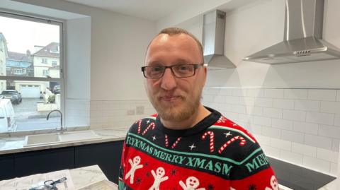 Andy Fletcher is pictured smiling at the camera and wearing a colourful Christmas jumper. He is wearing black glasses and is standing in one of the new kitchens. The kitchen has dark grey/blue units with white marble-effect surfaces and white tiles.