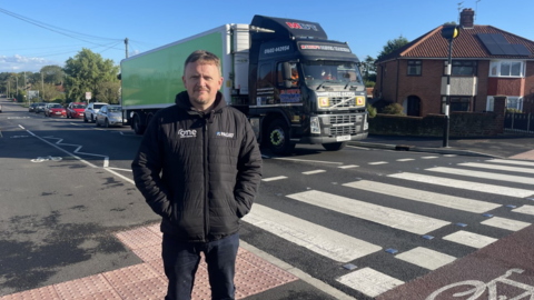 Matthew Moore in a black jacket standing beside a pedestrian crossing, as traffic passes in the background