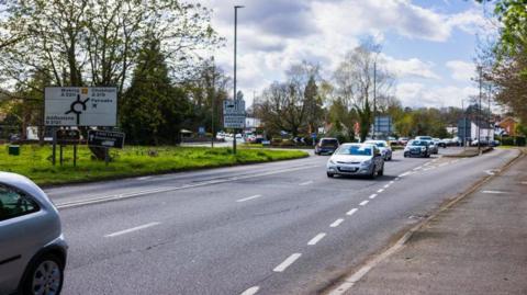 The image shows a road with around six cars driving towards the left. Grass can be seen on the left-hand-side of the road with a sign showing the different road directions. Some clouds can be seen in the sky, which is a pale blue.  Trees can be seen on both sides of the road.