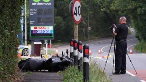 A forensic investigator, in a black shirt and trousers, taking a photograph of a black motorcycle which is lying on the ground. He is standing near a petrol station and there is a police car parked on the side. 