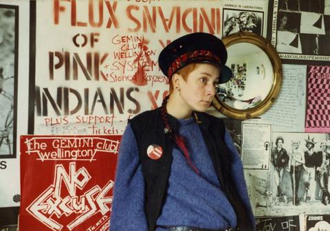 A woman stands against a wall covered with posters. The woman has very short red hair and she is wearing a blue jumper, a black waistcoat and a black cap with a red sash tied around it. The posters behind her have a mixture of writing, drawings photographs on them.