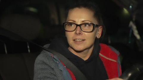 Shot of a woman sitting in the driver's seat of a car. She is wearing a grey jacket with a red trim and black-rimmed spectacles.