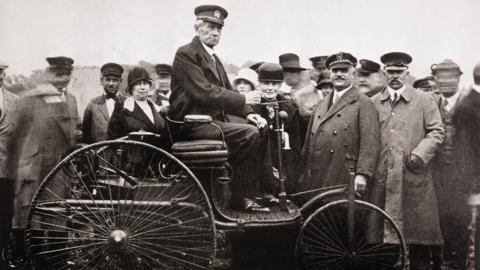 A black and white photo of Karl Benz displaying the Benz Patent-Motorwagen to a crowd of people. He is sat high up in the vehicle, which has large back wheels and a smaller front wheel, and is donning a suit and hat.