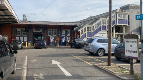 Twyford station car park showing cars in spaces, a welcome sign and people near a ticket machine by station entrance. Enclosed wooden stairs can be seen going up and over the rail line.