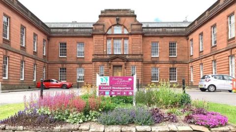 Dumfries and Galloway Council headquarters is a large sandstone building with rows of long white framed windows. The front of the building has threes sides with a car park and floral display in the middle.