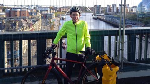 A man holding his cycle bike on a bridge with the river behind him, on either side and roads and buildings and clear skies behind.