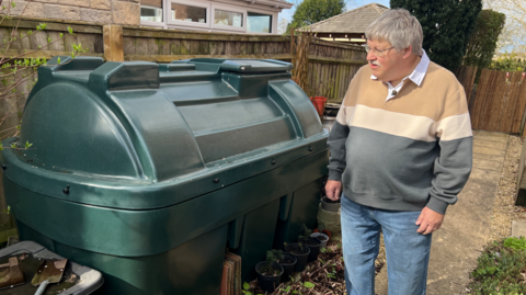 A man in a jumper stands next to a green oil tank