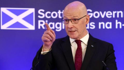 John Swinney gives a speech in Edinburgh. He is wearing a black suit jacket, a maroon tie and a white shirt.