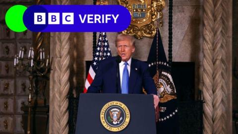 President Donald Trump speaks to reporters from a lectern bearing the seal of the US president. An American flag and a flag bearing the president's seal are behind him.  He wears a dark suit and a blue tie. 