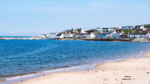 Portmahomack is a coastal village with white-walled houses and buildings related to the fishing industry and its harbour. In the foreground is a sandy beach. The picture was taken on a sunny day and the sky is blue and cloudless.