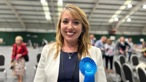 A smiling woman with long blonde hair wearing a white blazer, a navy top and a blue rosette standing in a busy hall.