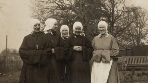 Five women stand together, facing the camera and smiling in a sepia-toned photograph. Behind them is a large tree with a wooden bench beneath it. Four of the women are wearing the black habit clothing of nuns, with white headpieces. The woman on the far left has a large crucifix hanging from her neck in front of her clothes. The woman on the far right is wearing a lighter coloured robe with a white pinafore tied around her waist and a white headpiece.