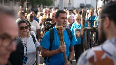 A young man carrying a gold baton - similar to the one containing the Olympic flame - through a crowd of people. He has short dark hair, an expression of concentration and is wearing short sleeved blue T-shirt and also carrying a black back pack.