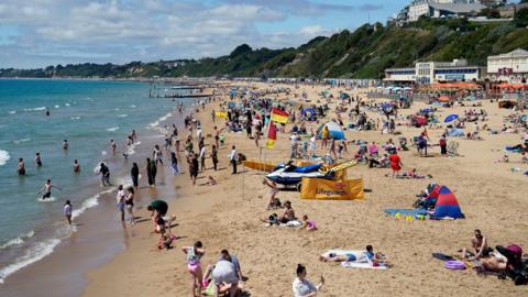 The beach under West Cliff, Bournemouth. The sand and sea are packed with families enjoying the sunshine.