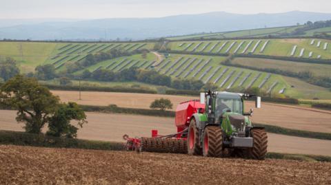 A green and red tractor is pulling a cultivator and drill across a muddy field. On the hills in the background lie solar panels. A light mist sits in the sky.